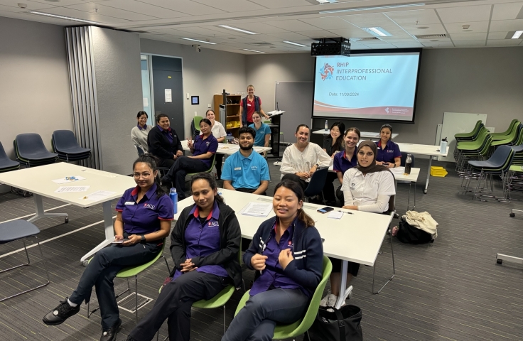 The image shows a classroom setting with individuals seated at tables arranged in a U-shape. At the front, a presentation screen displays the text "RHIP Interprofessional Education” and the date “11/09/2024.” A person stands to the right of the screen, addressing the attendees. 